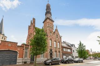 Achter deze monumentale gevel schuilt een herenwoning die haar geschiedenis moeiteloos combineert met een verrassende hoeveelheid leefruimte. Het herenhuis, ontworpen door architect Edward Careels, opgetrokken in <strong>neo-Vlaamserenaissancestijl en gedateerd 1892, </strong>werd als beschermd monument erkend om zijn artistieke waarde en onderscheidt zich door zijn uitgesproken architectuur, royale vertrekken en karaktervolle details. Achteraan vormt de bijzonder groene tuin met zwembad een onverwacht rustig decor bij dit statige geheel.<br /><br />De ligging in het hart van Zandhoven maakt het plaatje compleet: winkels, bakker, horeca, openbaar vervoer en dagelijkse voorzieningen bevinden zich op korte afstand, waardoor heel wat verplaatsingen eenvoudig te voet of met de fiets kunnen. Tegelijk zorgen de nabijgelegen E313 en E34 voor een vlotte verbinding richting Antwerpen en de omliggende gemeenten. Zo combineert deze locatie het gemak van wonen in het dorpscentrum met een bijzonder aangename private buitenruimte achter de woning.<br /><br /><strong>Indeling</strong><br />De statige inkomhal vormt het centrale vertrekpunt van de woning en geeft toegang tot de verschillende leefruimtes. Aan de straatzijde bevindt zich de bijzonder royale woonkamer met eetruimte, die achteraan verderloopt naar de lichtrijke veranda en zo een mooie verbinding maakt met de tuin.<br />Verder beschikt het gelijkvloers over een afzonderlijke eetkamer aansluitend de keuken en wasplaats, maar ook over een bureau met aanpalend archief. Die extra vertrekken maken de woning bijzonder interessant voor wie wonen en werken onder één dak wil combineren. Via een hall bereikt u bovendien een ruime hobbyruimte/extra bureel, de technische ruimte van het zwembad en de inpandige garage.<br /><br />De tuin vormt zonder twijfel één van de verrassingen van deze woning. Achter de historische gevel ontvouwt zich een verzorgde, groene buitenruimte met ruime terrassen, pergola en zwembad, omgeven door volwassen beplanting die voor een aangename besloten sfeer zorgt. Een automatische poort achteraan het perceel biedt rechtstreeks toegang tot de tuin en zorgt voor extra praktisch gemak.<br /><br />Op de <strong>eerste verdieping</strong> bevinden zich 3 volwaardige slaapkamers, een dressing en twee badkamers.<br /><br />De <strong>tweede en derde verdieping vormen samen een bijzonder ruim duplexgedeelte</strong> met een grote woonkamer, eigen keuken en badkamer, een karaktervolle torenkamer en op het bovenste niveau nog een slaapkamer. Hierdoor ontstaat een bijna zelfstandige woonverdieping die zich uitstekend leent als gastenverblijf, thuiskantoor of eigen plek voor opgroeiende kinderen en tieners.<br />Ook aan bergruimte ontbreekt het niet: de woning beschikt over een ruime wijnkelder en tal van bijkomende functionele ruimtes.<br /><br /><strong>Bijzonderheden</strong><br />Met haar 20 zonnepanelen, uitzonderlijke oppervlakte, vier slaapkamers, verschillende leef- en werkruimtes, inpandige garage, zwembad en afzonderlijk bruikbaar duplexgedeelte biedt deze woning een flexibiliteit die zelden voorkomt. Vooral de combinatie van de historische gevel, de besloten groene tuin en de verrassend veelzijdige indeling maakt het geheel bijzonder.