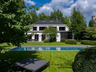<p><span>Deze karaktervolle Vlassak-Verhulst-villa biedt een uitzonderlijke verbinding tussen architectuur, wooncomfort en natuur. De woning is prachtig gelegen in de gemeente Zoersel, op de grens met Schilde, en grenst rechtstreeks aan het indrukwekkende Driehoeksbos. De aangelegde tuin vormt hierdoor een natuurlijk verlengstuk van de leefruimtes.</span></p>
<p><span>Binnen geniet u van royale en lichtrijke leefruimtes, waarbij de open woonkamer en eetruimte via grote raampartijen rechtstreeks in verbinding staan met de tuin en terrassen. De keuken met aangrenzende tuinkamer is ideaal voor wie graag binnen en buiten laat samenvloeien.</span></p>
<p><span>Een absolute troef is de uitgebreide poolzone met zwembad op basis van zoutelektrolyse. Het ruime poolhouse beschikt over een volledig uitgeruste buitenkeuken met Braai Master, Big Green Egg, vaatwasser en koelkast. Daarnaast zijn er een sauna, buitendouche, irrigatiesysteem en grondwaterput. Een ruime fietsenberging biedt bijkomende praktische bergruimte en herbergt tevens de zwembadinstallatie.</span></p>
<p><span>Op de eerste verdieping bevinden zich vier slaapkamers. De master bedroom beschikt over een dressing en ensuite badkamer. Daarnaast zijn er nog twee bijkomende badkamers en een zolderruimte.</span></p>
<p><span>Ook op het gelijkvloers werd aandacht besteed aan comfort en functionaliteit, met een ruime inkomhal met gastentoilet en vestiaire, een praktische berging met maatkasten (voormalige garage) en een kelder.</span></p>
<p><span>De woning is uitstekend onderhouden en afgewerkt met hoogwaardige materialen zoals parket, natuursteen en maatwerkinterieurs. Het comfort wordt verder aangevuld met airconditioning, een alarmsysteem en centrale verwarming. De woning beschikt over een gunstig EPC-label B.</span></p>
<p><span>Buiten geniet u van een prachtig aangelegde, zuidgerichte tuin met volwassen groen, verschillende terrassen, optimale privacy en meerdere autostaanplaatsen.</span></p>
<p><span>Een unieke eigendom voor wie op zoek is naar ruimte, rust, privacy en een hoogwaardige woonomgeving, met het Driehoeksbos letterlijk als achtertuin.</span></p>
<p><span>Ontdek deze bijzondere villa tijdens een persoonlijke bezichtiging.</span></p>