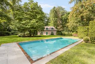 Verscholen in het groen van de gegeerde Bellevuedreef bevindt zich deze karaktervolle klassieke villa op een indrukwekkend parkdomein van ca. 1,4 hectare. Een eigendom waar ruimte, privacy en een bijzonder aangename woonbeleving samenkomen, omgeven door een volgroeide en zorgvuldig aangelegde tuin.<br /><br />De villa beschikt over ca. 575 m² bewoonbare oppervlakte en wordt aangevuld met een volwaardig gastenverblijf in het poolhouse, een afzonderlijke conciërgewoning, een inpandige garage voor drie wagens en een verwarmd buitenzwembad. De omvang van het domein, de volwassen begroeiing en de verschillende woon- en verblijfsfuncties maken van deze eigendom een bijzonder compleet geheel op een van de meest residentiële locaties van Schilde.<br /><br />Indeling gelijkvloers:<br />De centrale inkomhal met vestiaire en gastentoilet vormt het vertrekpunt van de woning en geeft toegang tot de verschillende leefruimtes.<br />De royale leefruimte omvat een zitkamer en eetkamer, aansluitend op een afzonderlijke studeerkamer. Daarnaast bevindt zich een ruim bureel met maatwerkkasten, waardoor ook professioneel of rustig thuiswerken perfect in de woning kan worden geïntegreerd.<br />Centraal in het dagelijkse leven staat de ruime leefkeuken, opgebouwd rond een gezellige ontbijtruimte met grote schuiframen naar het terras en de tuin. Het keukengedeelte met eiland is uitgerust met diverse inbouwtoestellen. Een praktische berging zorgt voor een tweede verbinding met de eetkamer, terwijl de bijkeuken met afzonderlijke zij-ingang bijkomende functionaliteit biedt.<br /><br />Eerste verdieping:<br />De nachthal leidt naar vier ruime slaapkamers, die elk over een eigen badkamer beschikken.<br />De mastersuite vormt een royaal privégedeelte met slaapkamer, dressing en badkamer. Daarnaast bevinden zich op deze verdieping een tweede ruime slaapkamer, een gastenkamer en een kinderkamer, telkens met een eigen badkamer.<br /><br />Vanuit de mastersuite is er via een tussengang en diensttrap verbinding met het gedeelte boven de garage.<br /><br />Conciërgewoning:<br />Boven de inpandige garage bevindt zich een afzonderlijk verblijfs- en conciërgegedeelte, bestaande uit een polyvalente leefruimte, slaapkamer en badkamer. Dit gedeelte leent zich uitstekend voor inwonend personeel, een au pair, familie of gasten.<br /><br />Kelder:<br />Via een natuurstenen trap is de kelderverdieping bereikbaar. Hier bevinden zich bijkomende bergruimtes, de technische installaties voor de woning en het zwembad en een sfeervolle wijnkelder.<br /><br />Poolhouse & gastenverblijf:<br />Aan het verwarmde zwembad bevindt zich een royaal bijgebouw, architecturaal afgestemd op de hoofdwoning. Het poolhouse werd ingericht als een volwaardig zelfstandig gastenverblijf met woonkamer, keuken, slaapkamer en badkamer.<br />Samen met de omliggende terrassen en het zwembad ontstaat hier een bijzonder aangename omgeving voor ontspanning, familie en gasten, met een grote mate van privacy ten opzichte van de hoofdwoning.<br /><br />Bijzonderheden:<br />- Uitzonderlijk parkdomein van ca. 14.433 m²<br />- Volledig omheind domein met maximale privacy<br />- Volgroeide tuin met uitgestrekte gazons en volwassen bomen<br />- Regelmatig onderhouden en verder opgefrist<br />- Verwarmd buitenzwembad<br />- Volwaardig poolhouse ingericht als zelfstandig gastenverblijf<br />- Afzonderlijk conciërge- of personeelsverblijf<br />- Inpandige garage voor drie wagens<br />- Verschillende ruime terrassen rondom de woning<br />- Uitgebreide tuinverlichting<br />- Alarm- en camerabewaking<br /><br />Een bijzonder compleet residentieel domein waar ruimte, discretie en groen centraal staan, op een van de meest gewaardeerde locaties van Schilde.