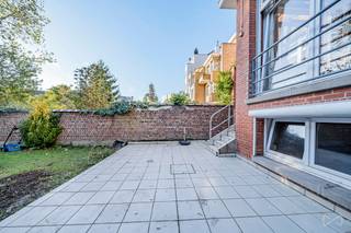 Gelegen in een rustige en residentiële wijk biedt dit gelijkvloers appartement met een oppervlakte van ongeveer 89 m² een aangename en comfortabele woonomgeving. Het bestaat uit een inkomhal, een ruime en lichte woon- en eetkamer, evenals een volledig uitgeruste keuken. Het appartement beschikt momenteel over één slaapkamer, maar een grote ruimte met toegang tot de tuin maakt het mogelijk om eenvoudig een tweede slaapkamer te creëren indien gewenst. Verder is er een douchekamer en een kelder aanwezig. Buiten vindt u een groot terras en een privétuin, ideaal om van zonnige dagen te genieten. Het pand is voorzien van een veiligheidsdeur, dubbele beglazing en nieuwe rolluiken aan alle ramen. Gelegen in een klein gebouw met lage lasten combineert dit appartement comfort, rust en nabijheid van winkels, openbaar vervoer en andere voorzieningen. Zeker een bezoek waard.