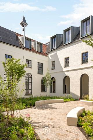 Binnen het Karmelproject bevindt zich een van de meest bijzondere woonentiteiten van het domein: de Kapelloft. Gevestigd in de voormalige neogotische kloosterkapel uit 1877, biedt deze eigendom een woonervaring die zelden voorkomt op de vastgoedmarkt.<br /><br />Bij de herbestemming van de kapel werd gekozen voor een doordacht box-in-the-box-concept, waarbij de oorspronkelijke kapelstructuur maximaal behouden blijft. Hierdoor blijven de indrukwekkende volumes, de hoge gewelven, de spitsboogvensters en het historische karakter van de ruimte volledig beleefbaar.<br /><br />De leefruimte bevindt zich centraal in de kapel en wordt gekenmerkt door een uitzonderlijk ruimtegevoel. Vanuit de keuken biedt een groot raam zicht op de uitgestrekte parktuin van het domein. Een draaitrap leidt naar een hoger gelegen bureau- of polyvalente ruimte die uitkijkt over het geheel.<br /><br />Aan de andere zijde van de loft bevindt zich een afzonderlijke woonmodule. Op het gelijkvloers zijn de master bedroom en de ensuite badkamer ondergebracht. Op de verdieping bevinden zich nog twee bijkomende slaapkamers en een tweede badkamer.<br /><br />Naast de privatieve woonruimtes genieten bewoners van toegang tot de gemeenschappelijke kloostertuin van circa 5.000 m². Met haar historische elementen, wandelpaden en volwassen groen vormt deze tuin een uitzonderlijke plek van rust midden in het historische centrum van Brugge.<br /><br />De Kapelloft toont hoe erfgoed een nieuwe bestemming kan krijgen zonder zijn identiteit te verliezen. Een eigendom waar geschiedenis, architectuur en hedendaags wooncomfort samenkomen in een setting die nauwelijks een vergelijking kent.<br /><br />Een unieke woonkans voor wie op zoek is naar ruimte, karakter en rust op een van de meest bijzondere locaties van Brugge.
