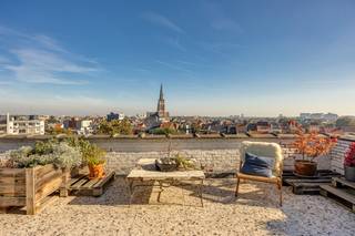 <strong>Algemeen</strong><br />Op de zesde en zevende verdieping, helemaal bovenaan het gebouw, ligt dit duplex-penthouse van ca. 211 m². Het voelt eerder als een huis in de lucht dan als een appartement. Beneden is het rustig, met drie slaapkamers, een aparte dressing, een badkamer en veel ingemaakte kasten. Boven ligt één grote, lichte leefruimte met witgeschilderde balken, een industriële keuken, een vierde slaapkamer, een tweede badkamer en twee terrassen van samen zo’n 70 m². Een smeedijzeren wenteltrap verbindt de twee verdiepingen. Ook technisch zit het goed: het dak van het gebouw werd gerenoveerd, de verwarmingsketel is pas vernieuwd en het appartement heeft een EPC-label C. Er is een lift, en bij het appartement horen twee kelders.<br /><strong>Ligging</strong><br />Het penthouse ligt aan het begin van de Turnhoutsebaan, op het kruispunt met de Carnotstraat en de Kerkstraat. Het Centraal Station, het Astridplein en de Zoo liggen op een paar minuten wandelen. Winkels, bakkers, koffiebars en restaurants vind je in alle richtingen, en tram en bus stoppen vlakbij. Je woont hier in het hart van de stad, maar vanaf de bovenste verdieping hoor je de drukte nauwelijks en zie je vooral daken, kerktorens en veel lucht.<br /><strong>Indeling</strong><br />Zesde verdieping: de nachtverdieping<br />Je komt binnen in een ruime inkomhal met een marmeren vloer en een hele wand ingemaakte kasten, zodat jassen, schoenen en tassen meteen uit het zicht zijn. Vanuit de hal bereik je alle ruimtes op deze verdieping.<br />Op deze verdieping liggen drie slaapkamers. De grootste is een echt ruime kamer met een groot raam en een deur naar het balkon, vanwaar je over de daken en het groen van de buurt kijkt. De tweede slaapkamer is nu ingericht als bureau. Een groot raam zorgt voor veel daglicht en de witgeschilderde bakstenen muur geeft de kamer karakter. De derde slaapkamer is even rustig en ruim, en elke kamer heeft eigen kastruimte. Daarnaast is er een aparte dressing met een eigen raam, dat uitkijkt op de kerktoren. Zo heb je kleding en schoenen netjes uit de slaapkamers. Op de hele verdieping, behalve in de inkomhal, ligt eiken mozaïekparket. De korte stroken liggen per vier in vierkantjes, telkens een kwartslag gedraaid zoals een dambord. Het is een klassieker die mooi verweerd is en de kamers meteen warmte geeft.<br />De badkamer op deze verdieping is sober en strak. De douche heeft grote antracietkleurige tegels, een regendouche met handdouche en een glazen schuifdeur. Het aparte toilet ligt ernaast.<br />De wenteltrap<br />Vanuit de inkomhal loop je via een smeedijzeren wenteltrap naar boven. Wie omhoog kijkt, ziet een ronde trapopening met een sierlijke balustrade vol krullen en daarboven de witte balken van de leefruimte. Het is een van die details die je nergens anders tegenkomt.<br />Zevende verdieping: de leefverdieping<br />Boven kom je uit in het hart van de woning: één grote leefruimte van meer dan twaalf meter lang. Het plafond heeft witgeschilderde houten balken en aan meerdere kanten zijn er ramen, waardoor er van ‘s ochtends tot ‘s avonds licht binnenvalt. De ronde trapopening staat midden in de ruimte en verdeelt ze vanzelf in zones zonder dat er muren nodig zijn. Aan de ene kant is er plaats voor een grote zithoek, met een witte nis in de muur en een stuk witgeschilderde baksteen die er een gezellige hoek van maken. Aan de andere kant staat de eettafel voor een groot raam met houten schuiframen, die rechtstreeks uitkomen op het terras.<br />De keuken is industrieel ingericht: witte metrotegels tot aan het plafond, een stoer professioneel fornuis met grote oven, een roestvrijstalen dampkap met zichtbare afvoerbuis, open rekken in staal en hout en een magneetstrip voor de messen. De dubbele spoelbak staat onder een breed raam, zodat je bij de afwas over de stad kijkt. Het is een keuken voor mensen die graag en veel koken.<br />Dan de terrassen. Het zuidwestterras van ca. 40 m² krijgt de zon in de namiddag en ‘s avonds. Het noordoostterras van ca. 30 m² is ideaal voor een ontbijt in de ochtendzon of als koele plek op warme zomerdagen. Op het terras liggen terrazzotegels en rondom staat een witgeschilderde bakstenen borstwering. Het uitzicht reikt over de daken tot aan de skyline van Antwerpen.<br />Op deze verdieping ligt ook de vierde slaapkamer, een rustige kamer met sierlijsten aan het plafond. De tweede badkamer is een echte eyecatcher. De wanden zijn bekleed met handgemaakte, glanzende tegeltjes in zachte grijsblauwe tinten, en er is een ligbad, een inloopdouche met regendouche, een lange brede wastafel met een grote spiegelkast en een handdoekradiator. Door het raam valt daglicht binnen. Verder is er een apart toilet en een berging die als wasplaats is ingericht.<br />De hele bovenverdieping heeft een bamboevloer met de typische knopen in het hout, die een ander karakter heeft dan het parket beneden. In de leefruimte zijn de vloerconvectoren weggewerkt onder houten lattenroosters, zodat er geen radiatoren tegen de muren staan.<br /><strong>Troeven</strong>
<ul>
<li>Duplex-penthouse van ca. 211 m² op de 6e en 7e verdieping</li>
<li>Twee terrassen: zuidwest (ca. 40 m²) en noordoost (ca. 30 m²), met weids zicht over Antwerpen</li>
<li>Leefruimte van ruim 2 m1 met witgeschilderde balken en veel lichtinval</li>
<li>Industriële keuken met professioneel fornuis</li>
<li>4 ruime slaapkamers (3 op de 6e, 1 op de 7e), een dressing en veel ingemaakte kasten</li>
<li>2 volwaardige badkamers (één per verdieping) en 2 aparte toiletten</li>
<li>Smeedijzeren wenteltrap als blikvanger</li>
<li>Eiken mozaïekparket op de 6e verdieping, bamboe op de 7e, marmer in de inkomhal</li>
<li>Vloerconvectoren in de leefruimte</li>
<li>Gerenoveerd dak, nieuwe verwarmingsketel, EPC C</li>
<li>Lift en 2 kelders</li>
<li>Op wandelafstand van het Centraal Station</li>
</ul>
De grondplannen, het EPC en de asbestinventaris vind je onder “Documenten” op onze website. Kom het uitzicht zelf bekijken en maak een afspraak via info@wellimmo.net of op 03 225 00 68.