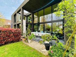Bent u op zoek naar een unieke woonst waar licht, ruimte en privacy samenkomen? Lees dan zeker verder!Deze ruime open villa van 15 x 15 meter is gelegen op een prachtig perceel van 1.132 m² in een idyllische, landelijke omgeving. Met een gunstig EPC-label B voldoet de woning ruimschoots aan de huidige energetische normen. De volgroeide, aangelegde tuin biedt volop privacy, terwijl de N16 en E17 zorgen voor een uitstekende bereikbaarheid.Bij aankomst springt de indrukwekkende voorgevel meteen in het oog. De imposante voordeur van 5,35 meter hoog en 2,20 meter breed bevindt zich op 16 meter van de straatkant en is strategisch verscholen in een lichtkoker achter een volle muur. Een slim geplaatst raam van 1 x 6 meter voorkomt inkijk en biedt vanuit de woning een panoramisch zicht op de groene voortuin. Binnen zorgen de open indeling en twee vides voor een uitzonderlijk ruimtegevoel. De leefruimte van 12 x 6 meter heeft een plafondhoogte van 5,35 meter en wordt overspoeld door daglicht dankzij de indrukwekkende raampartijen. De 3 meter diepe oversteek van zijgevel en dak fungeert als natuurlijke zonwering, waardoor de woning ook op warme dagen aangenaam koel blijft. De open leefkeuken beschikt over een groot keukeneiland, ruime kasten en grote plafondhoge ramen. Op het gelijkvloers loopt de authentieke Belgische blauwe hardsteen drempelloos door van de inkomhal tot het terras, waardoor binnen en buiten één geheel vormen.De bovenverdieping straalt warmte uit dankzij de Vlaamse eiken parketvloer. De master bedroom sluit aan op een indrukwekkende glaswand van 15 meter lang en 2,55 meter hoog, gevolgd door een dressing en badkamer met dubbele lavabo, toilet en ruime inloopregendouche. Een praktische wasberging en comfortabele logeerkamer vervolledigen deze verdieping. U hoeft dus nooit meer met zware wasmanden de trap op en af! De woning is uitgerust met lage-temperatuurvloerverwarming via een hoogwaardige Weishaupt-condensatieketel. Twee afzonderlijke circuits en thermostaten laten toe om boven en beneden onafhankelijk te verwarmen.Ook de tuin is een absolute troef, met 29 volwassen bomen, diverse hagen, fruitbomen en een walnotenboom. De regenwaterput van 10.000 liter is aangesloten op twee toiletten en kan ook op de wasmachine worden aangesloten. De dubbele inpandige garage biedt plaats aan twee wagens naast elkaar.Wilt u zelf de unieke sfeer, ruimte en architectuur van deze villa ervaren? Neem dan zeker contact met ons op voor een bezoek!