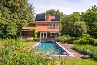 <p>In een van de meest gegeerde residentiële buurten van Brasschaat, midden in het groene Ter Borcht, bevindt zich deze karaktervolle villa op een prachtig aangelegd perceel van ca. <strong>4.850 m²</strong>. Omringd door volwassen bomen en een uitzonderlijk mooie tuin geniet u hier van absolute rust en privacy.</p>
<p>De woning, oorspronkelijk gebouwd in 1928, werd ongeveer achtien jaar geleden grondig gerenoveerd en sindsdien steeds met veel zorg onderhouden en verder gemoderniseerd. Zo werden recent nog een volledig nieuw dak, zonnepanelen en een laadpaal geplaatst.</p>
<p>Via de inkomhal met gastentoilet bereikt u het bureau, de garage en de leefruimtes. De royale woonkamer met parketvloer, open haard en gezellige zithoek baadt in het licht dankzij de grote raampartijen en biedt een prachtig uitzicht over de tuin en het zwembad.</p>
<p>De ruime leefkeuken, afgewerkt met natuursteen en voorzien van alle hedendaagse toestellen, vormt het hart van de woning en geeft rechtstreeks uit op het zonnige terras.</p>
<p>Op de eerste verdieping bevinden zich twee ruime slaapkamers, elk met een eigen badkamer. De tweede verdieping biedt nog twee bijkomende slaapkamers en een douchekamer, ideaal voor een groot gezin of gasten.</p>
<p>Buiten vormt de schitterend aangelegde tuin een ware oase van rust. Het verwarmde zwembad (14 x 4 m) met warmtepomp, de verschillende terrassen en de volwassen beplanting zorgen voor een uitzonderlijke woonbeleving in een groene omgeving.</p>