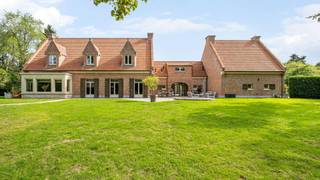 Verscholen op een prachtig perceel van maar liefst 35a 89ca ligt deze unieke villa, een eigendom die zich moeilijk laat vergelijken met het klassieke woningaanbod. Achter de poort ontvouwt zich een domein waar authentieke architectuur, royale volumes en een groene, volledig privatieve tuin naadloos in elkaar overvloeien. <br /><br />De villa werd in 1978 gebouwd door een aannemer die koos voor duurzame materialen en verfijnd vakmanschap. Vandaag is die kwaliteit nog steeds zichtbaar in de talrijke authentieke elementen die de woning haar ziel geven. Franse zandstenen bogen, een karaktervolle erker, massief eiken deuren, elegante trappen en warme parketvloeren zorgen voor een tijdloze uitstraling die doorheen de jaren met zorg werd behouden. <br /><br />Bij het binnenkomen in de hoofdwoning wordt u verwelkomd in een ruime inkomhal waar de indrukwekkende Franse zandstenen bogen meteen de toon zetten. De authentieke materialen en het rijke eiken schrijnwerk creëren een sfeer van klasse en warmte. Vanuit de inkomhal vloeit de woning verder naar de verschillende leefruimtes, bestaande uit een sfeervolle woonkamer, een gezellige eetkamer en een ruime keuken waar het dagelijkse leven zich ongetwijfeld afspeelt. Daarnaast beschikt het gelijkvloers over een bureau, ideaal voor thuiswerk of een vrij beroep, een praktische bijkeuken en extra bergruimte. <br /><br />Op de verdieping bevinden zich drie ruime slaapkamers en een badkamer. Via een zolderluik bereikt u de geïsoleerde zolder, die perfect dienst doet als extra opslagruimte. Ondergronds beschikt de woning bovendien over een kelder, wijnkelder en kruipkelder. <br /><br />Doorheen de woning zorgen de combinatie van natuursteen, massief eikenhout en parketvloeren voor een bijzonder warme sfeer. Hier voelt elke ruimte authentiek aan, zonder in te boeten aan comfort. <br /><br />Een absolute meerwaarde van deze eigendom is de volledig geïntegreerde tweede woongelegenheid. Via een charmante overdekte boogverbinding staat deze zelfstandig woonunit in contact met de hoofdwoning, terwijl beide delen toch hun eigen privacy behouden. <br /><br />Deze tweede woongelegenheid werd de afgelopen jaren grondig gemoderniseerd en straalt hedendaagse klasse uit. De lichtrijke leefruimte met open keuken vormt het centrale hart van de woning. De maatwerkkeuken met royaal kookeiland, geïntegreerde toestellen en uitgebreide kastruimte werd ontworpen met oog voor functionaliteit en esthetiek. Een elegante metalen wenteltrap leidt naar de verdieping, waar een ruime slaapkamer met ingebouwde kastenwand en een comfortabele badkamer met ligbad en aparte douche werden ingericht. <br /><br />Ook buiten onderscheidt deze eigendom zich moeiteloos. De prachtig onderhouden tuin biedt een uitzonderlijk gevoel van rust en privacy. Volwassen beplanting, uitgestrekte groenzones en verschillende terrassen zorgen ervoor dat u op elk moment van de dag een aangename plek in de zon of schaduw vindt. <br /><br />De tweede woongelegenheid beschikt over een eigen terras met zonneluifel en een afzonderlijke tuinzone. Beide tuingedeeltes worden op een sfeervolle manier met elkaar verbonden via een fontein, waardoor het geheel aanvoelt als een harmonieus privépark. <br /><br />Deze eigendom biedt een zeldzame combinatie van authentieke charme, kwalitatieve afwerking, uitzonderlijke oppervlakte en flexibele woonmogelijkheden. Een ideale thuis voor grote gezinnen, kangoeroewonen, zorgwonen of wie wonen en werken wenst te combineren in een unieke en stijlvolle omgeving.