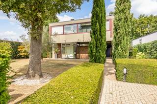 In een groene en rustige omgeving aan de rand van Hasselt te Genkersteenweg 290 bevindt zich deze bijzondere eigendom: een architecturale parel die stijl, ruimte en functionaliteit moeiteloos combineert. Dit is geen doorsnee woning, maar een statement – ontworpen met visie, gebouwd met vakmanschap en door de jaren heen zorgvuldig onderhouden. De villa werd getekend door de gerenommeerde architect Dolf Nivelle, wiens handtekening duidelijk herkenbaar is in de tijdloze vormgeving. Robuuste materialen, strakke lijnen en een optimaal gebruik van licht en ruimte vormen de rode draad doorheen het ontwerp. Het perceel van 9a 57ca is volledig omheind en beschikt over twee toegangswegen: voorkant (hoofdtoegang): een royale oprit met parkeermogelijkheden voor meerdere wagens, ideaal voor bewoners én bezoekers, achterkant (Sluisstraat): een discrete toegang, perfect voor professioneel gebruik of afzonderlijk bewonersverkeer. Deze dubbele bereikbaarheid maakt het pand bijzonder geschikt voor wie wonen en werken wil combineren. Bovenverdieping (woongedeelte): Een sfeervolle leefruimte met overvloedig daglicht, warme houten vloeren, een gezellige haard en een veranda met zicht op groen. De royale keuken sluit naadloos aan op de leefruimte. Verder zijn er drie comfortabele slaapkamers, een badkamer, gastentoilet en praktische berging. Benedenverdieping (professioneel gedeelte): Recent vernieuwde ruimtes met aparte inkom, wachtruimte, kitchenette en sanitaire voorzieningen. Ideaal als kantoor, praktijk, creatieve studio of consultatieruimte. Dankzij de flexibele indeling kan deze verdieping ook dienen als afzonderlijke woonunit (bv. kangoeroewonen, co-housing of zorgwoning). Authentieke materialen zoals natuursteen, hout en staal zorgen voor een krachtige en tijdloze uitstraling. De esthetische coherentie van de woning straalt pure klasse uit, zonder in te boeten aan hedendaags comfort. Fraai aangelegde tuin met maximale privacy, ruim zonneterras met zuidwestoriëntatie, carport en meerdere parkeermogelijkheden, aparte wellnesshoek met sauna – ideaal voor ontspanning na een drukke dag. Slechts enkele minuten van het bruisende centrum van Hasselt, vlak bij natuurgebieden, vijvers en bossen – perfect voor wandelaars en fietsers, optimale visibiliteit en toegankelijkheid voor professionele doeleinden, nabij Bokrijk, Kiewit en belangrijke invalswegen. Troeven: exclusieve architectuur van Dolf Nivelle, flexibele indeling met aparte toegangen voor privé en professioneel gebruik, professioneel ingerichte, gerenoveerde benedenverdieping, mogelijkheden voor vrij beroep, praktijk aan huis, co-housing of zorgwoning, uitstekend onderhouden en instapklaar, zuidwestgericht terras en aparte sauna-unit, ruime parkeergelegenheid, groen en rustig gelegen, toch vlak bij het stadscentrum. Kenmerken: bewoonbare opp.: 282 m², opp. perceel: 9a 57ca, EPC: 458 kWh/m² jaar, KI: € 1.899, bouwjaar: 1962 Een mooie kans voor zij die opzoek zijn naar immo te koop in Hasselt. Relevante documenten zijn eveneens beschikbaar op onze website www.dewaele.com of geef ons een seintje op 011 30 50 50. Dit pand is onderworpen aan de renovatieverplichting van de Vlaamse overheid voor residentiële gebouwen (behoudens bij sloop binnen 5 jaar na de aankoop). Consulteer voor meer informatie de website van het Vlaams Energie- en Klimaatagentschap via https://www.vlaanderen.be/een-woning-kopen/renovatieverplichting-voor-residentiele-gebouwen