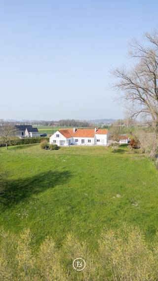 Ontdek deze charmante landelijke woning met prachtige vergezichten op een uitzonderlijk perceel van ca. 78 are, omgeven door uitgestrekte weilanden. Deze eigendom is uitermate geschikt voor kangoeroewonen of de combinatie van wonen en werken, dankzij de aparte bureauruimte en de volledig uitgeruste inpandige studio.<br /><br />Bij het betreden van de woning wordt u verwelkomd in de inkomhal met gastentoilet. De lichtrijke leefruimte vormt het hart van de woning en biedt een gezellige zithoek en eetplaats, beide met een prachtig zicht op de tuin en het omliggende landschap. Aansluitend vindt u de uitgeruste keuken met eetgedeelte, een praktische berging en een koele ruimte.<br />De woning beschikt over een polyvalente ruimte die perfect ingericht kan worden als bureau of praktijkruimte. Op het gelijkvloers bevindt zich eveneens de nachthal die toegang geeft tot de master bedroom met eigen badkamer, drie kinderkamers, een douchekamer en apart toilet.<br />Op de eerste verdieping bevindt zich de inpandige studio, voorzien van een leefruimte, keuken, slaapkamer en badkamer, ideaal voor inwonende familie. Het tweede deel van deze verdieping biedt bovendien nog tal van uitbreidingsmogelijkheden.<br /><br />De buitenomgeving is zonder twijfel een absolute meerwaarde: een zuidgericht terras en een uitgestrekte tuin met weide creëren een oase van rust en ruimte, met prachtige vergezichten over het omliggend groen.