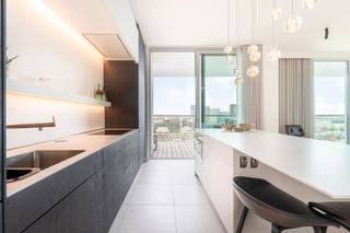 <p>Stijlvol appartement gelegen op het Eilandje, in het iconische Chipperfield-project, met spectaculair zicht. Gelegen op de 13e verdieping van een residentie met 15 verdiepingen, geniet u hier van een uitzonderlijk panoramisch uitzicht over de stad, de haven en het water.</p>
<p>Een brede, lichtrijke inkomhal biedt toegang tot alle kamers en leidt naadloos naar een ruime leefruimte. Een hoogwaardige Bulthaupt-keuken is voorzien van Miele-apparatuur, inclusief stoom- en grilloven. De leefruimte, met 3,2 meter hoge plafonds, is afgewerkt met een warme parketvloer en beschikt over ingemaakt kastenwerk met bar en een elegante ethanolhaard. Dankzij de kamerhoge raampartijen en meerdere glazen schuifpuien geniet u vanuit elke hoek van een adembenemend panorama over het Kattendijkdok en de Antwerpse skyline.</p>
<p>Vanuit alle kamers heeft u toegang tot het zuidgerichte omloop-hoekterras, één van de unieke troeven van dit appartement. Op deze hoogte geniet u ongestoord van het water, de stad en de haven — met een glazen balustrade die het zicht maximaal vrijlaat.</p>
<p>Verder beschikt het appartement over een master-slaapkamer met ensuite badkamer voorzien van inloopdouche, dubbele wastafel en toilet, afgewerkt in Corian. De oorspronkelijke tweede doucheruimte werd omgebouwd tot een praktische wasplaats (volledig ingericht, maar eenvoudig opnieuw als douchekamer te gebruiken). Daarnaast is er een tweede kamer ingericht als bureau met ingebouwde kasten en een geïntegreerd stapelbed, waardoor deze ook perfect kan functioneren als volwaardige tweede slaapkamer.<br /><br /><strong>De twee ondergrondse autostaanplaatsen kunnen aangekocht worden voor € 80.000; de kelderberging is inbegrepen.</strong></p>
<p>Een zeldzame kans om te wonen op één van de meest exclusieve locaties van Antwerpen, hoog boven de stad, in één van de meest gegeerde en dynamische buurten waar erfgoed en hedendaagse architectuur elkaar ontmoeten. Op wandelafstand van het MAS, sfeervolle terrassen aan het water, toprestaurants, boetieks en uitstekende verbindingen met de rest van de stad.</p>
