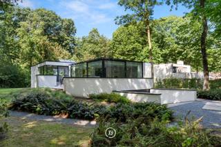 Deze moderne villa uit 2018 is discreet gelegen op een mooi perceel van 2.461 m². De woning en tuin liggen dieper op het perceel, bereikbaar via een lange oprit en afgesloten door een automatische poort. Hierdoor ontstaat een bijzonder private woonomgeving, omgeven door groen en volwassen bomen.<br /><br />De strakke architectuur werd ontworpen met veel aandacht voor licht, ruimte en het contact met de omgeving. De royale leef- en eetruimte bevinden zich op het gelijkvloers en bieden rondom zicht op het groen. Ook de keuken sluit hier mooi op aan en beschikt over een ruime en praktische keukenberging. Het interieur werd bovendien recent volledig herschilderd.<br /><br />De woning telt 340 m² bewoonbare oppervlakte en beschikt over vier slaapkamers, waarvan één momenteel als bureel is ingericht. De ruime master suite beschikt over een eigen ensuite badkamer, terwijl voor de overige kamers een afzonderlijke douchekamer voorzien is.<br /><br />De slaapkamers, badkamers, berging, ruime garage, technische ruimte en wasruimte bevinden zich gedeeltelijk onder het maaiveld. Deze bijzondere indeling zorgt ervoor dat de leefruimtes maximaal van het daglicht en de groene omgeving genieten, terwijl de lagergelegen ruimtes ook tijdens warme zomerdagen aangenaam koel blijven.<br /><br />Ook energetisch is de woning volledig mee met haar tijd, met zonnepanelen, een E-peil van E54 en EPC-label A. Een hedendaagse villa waar strakke architectuur, privacy, groen en modern wooncomfort op een serene manier samenkomen.