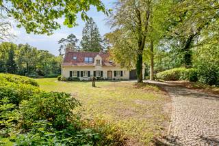 Op een discreet perceel treffen we deze karaktervolle villa in landelijke architectuur. De woning is omgeven door een volwassen tuin met imposante bomen en geniet hierdoor van een aangename privacy.<br /><br />Binnen valt meteen het warme karakter van de woning op. De royale leefruimte wordt gekenmerkt door zichtbare houten balken, een terracottavloer, een natuurstenen open haard en talrijke raampartijen die voor veel natuurlijk licht en een mooie verbinding met de tuin zorgen. De hedendaagse leefkeuken vormt samen met de afzonderlijke ontbijthoek een aangename dagelijkse leefplek. Met 3 a 4 slaapkamers, twee badkamers, een ruim bureau, een inpandige garage en een volwaardige master suite biedt deze woning bovendien een bijzonder comfortabele gezinsindeling.<br /><br />Indeling gelijkvloers:<br />De inkomhal geeft toegang tot de verschillende vertrekken op het gelijkvloers. Een afzonderlijk en lichtrijk bureau, voorzien van brede raampartijen met zicht op het groen, biedt een aangename thuiswerkplek en kan tevens polyvalent worden ingericht.<br /><br />Centraal in de woning bevindt zich de ruime leefkamer met zit- en eetgedeelte. De houten balkenstructuur, terracottavloer en natuurstenen open haard zorgen hier voor een warme en karaktervolle uitstraling. Grote ramen en tuindeuren versterken het contact met de omliggende tuin. De open trap en vide geven het geheel bovendien een aangenaam gevoel van ruimte.<br /><br />De leefkeuken werd hedendaags ingericht met een royaal centraal kookeiland, veel kastruimte en kwalitatieve toestellen. Aansluitend bevindt zich een afzonderlijke ontbijthoek met veel natuurlijke lichtinval.<br /><br />Verder beschikt het gelijkvloers over een inpandige garage die eveneens plaats biedt aan de wasvoorzieningen en bijkomende bergruimte.<br /><br />Eerste verdieping:<br />De eerste verdieping omvat een volwaardig slaapgedeelte met in totaal 3 slaapkamers en een dressing die ook als 4e slaapkamer kan dienen.<br /><br />Een ruime hoofdslaapkamer, met hiernaast een dressing met maatkasten en hiernaast een badkamer. Deze badkamer is uitgerust met een dubbele wastafel, ligbad en afzonderlijke douche.<br /><br />Daarnaast bevinden zich op deze verdieping nog 2 bijkomende slaapkamers, geschikt als kinder-, logeer- of hobbykamers en een ruime bureauhoek op de overloop. Zij beschikken over een 2e badkamer met ligbad, wastafel en toilet.<br /><br />Kelder:<br />Onder een gedeelte van de leefruimte bevindt zich een praktische / wijn- kelder, bereikbaar via een luik in de vloer, eveneens geschikt als bijkomende voorraad- of bergruimte.<br /><br />Tuin:<br />De woning is mooi ingeplant op een ruim en volwassen aangelegd perceel. Grote gazonpartijen, volgroeide bomen, hagen en groenmassieven zorgen voor een bijzonder aangenaam kader en een hoge mate van privacy. De kasseiverharding en landelijke architectuur sluiten harmonieus aan bij het groene karakter van de eigendom.<br /><br />Bijzonderheden:<br />- Perceel van ca. 3.743 m² volgens opmetingsplan<br />- Nuttige oppervlakte volgens kadaster ca. 326 m²<br />- 3 à 4 slpks<br />- 2 badkamers<br />- Ruim afzonderlijk bureau<br />- Asbestveilig<br />- P-score A – G-score A