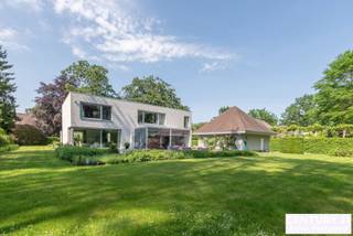 <div>
<div>
<section>
<div>
<div>
<div>
<div>
<div>
<div>
<h2>Exclusive Architect-Designed Villa in a Prime Location in De Lint Residential Estate</h2>
<p>Set on a beautiful southwest-facing plot within walking distance of the unique De Liereman nature reserve, this contemporary architect-designed villa offers an exceptional blend of timeless architecture, generous living spaces and high-quality finishes. Ideally located, both Eindhoven and Antwerp are only approximately 25 minutes away.</p>
<p>Upon entering, you are welcomed by a spacious entrance hall with cloakroom and guest toilet. Elegant double doors lead to the impressive living room, where large windows frame panoramic views of the beautifully landscaped garden. The abundance of natural light and seamless connection with the outdoors create a truly exceptional living experience. Adjacent to the living area is an open space, ideal as a home office.</p>
<p>A stylish double-sided fireplace elegantly connects the living room with the spacious kitchen-diner. The kitchen is impeccably maintained and features quality appliances. Large sliding doors provide direct access to the terraces and garden. A practical utility room and separate laundry room are located behind the kitchen. The ground floor is further complemented by an integrated double garage with automatic door and passenger lift.</p>
<p>Upstairs, the generous landing leads to the impressive master suite with walk-in dressing room and luxurious bathroom. Two additional full-sized bedrooms share a bathroom, while a spacious study can easily be converted into a fourth bedroom and benefits from its own private terrace.</p>
<p>Outside, the mature, fully enclosed garden offers complete privacy and sun terraces and a covered terrace invite you to enjoy outdoor living. An additional detached garage building provides ample space for extra vehicles.</p>
<p>The villa is also exceptionally low-maintenance thanks to its durable aluminium window frames and high-quality brick façades, eliminating the need for exterior painting while preserving its elegant appearance.</p>
<h3>Key Features</h3>
<ul>
<li>Southwest-facing plot with optimal sun exposure</li>
<li>Passenger lift to the first floor</li>
<li>Floor heating on both ground and first floors</li>
<li>Integrated double garage with automatic door</li>
<li>Detached garage building with storage loft</li>
<li>Alarm system</li>
<li>Exterior sun screens</li>
<li>Beautifully landscaped, fully enclosed garden</li>
<li>Large terraces and a covered, heated terrace</li>
</ul>
</div>
</div>
</div>
</div>
</div>
</div>
</section>
</div>
</div>