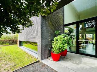 Bent u op zoek naar een unieke woonst waar licht, ruimte en privacy samenkomen? Lees dan zeker verder!Deze ruime open villa van 15 x 15 meter is gelegen op een prachtig perceel van 1.132 m² in een idyllische, landelijke omgeving. Met een gunstig EPC-label B voldoet de woning ruimschoots aan de huidige energetische normen. De volgroeide, aangelegde tuin biedt volop privacy, terwijl de N16 en E17 zorgen voor een uitstekende bereikbaarheid.Bij aankomst springt de indrukwekkende voorgevel meteen in het oog. De imposante voordeur van 5,35 meter hoog en 2,20 meter breed bevindt zich op 16 meter van de straatkant en is strategisch verscholen in een lichtkoker achter een volle muur. Een slim geplaatst raam van 1 x 6 meter voorkomt inkijk en biedt vanuit de woning een panoramisch zicht op de groene voortuin. Binnen zorgen de open indeling en twee vides voor een uitzonderlijk ruimtegevoel. De leefruimte van 12 x 6 meter heeft een plafondhoogte van 5,35 meter en wordt overspoeld door daglicht dankzij de indrukwekkende raampartijen. De 3 meter diepe oversteek van zijgevel en dak fungeert als natuurlijke zonwering, waardoor de woning ook op warme dagen aangenaam koel blijft. De open leefkeuken beschikt over een groot keukeneiland, ruime kasten en grote plafondhoge ramen. Op het gelijkvloers loopt de authentieke Belgische blauwe hardsteen drempelloos door van de inkomhal tot het terras, waardoor binnen en buiten één geheel vormen.De bovenverdieping straalt warmte uit dankzij de Vlaamse eiken parketvloer. De master bedroom sluit aan op een indrukwekkende glaswand van 15 meter lang en 2,55 meter hoog, gevolgd door een dressing en badkamer met dubbele lavabo, toilet en ruime inloopregendouche. Een praktische wasberging en comfortabele logeerkamer vervolledigen deze verdieping. U hoeft dus nooit meer met zware wasmanden de trap op en af! De woning is uitgerust met lage-temperatuurvloerverwarming via een hoogwaardige Weishaupt-condensatieketel. Twee afzonderlijke circuits en thermostaten laten toe om boven en beneden onafhankelijk te verwarmen.Ook de tuin is een absolute troef, met 29 volwassen bomen, diverse hagen, fruitbomen en een walnotenboom. De regenwaterput van 10.000 liter is aangesloten op twee toiletten en kan ook op de wasmachine worden aangesloten. De dubbele inpandige garage biedt plaats aan twee wagens naast elkaar.Wilt u zelf de unieke sfeer, ruimte en architectuur van deze villa ervaren? Neem dan zeker contact met ons op voor een bezoek!