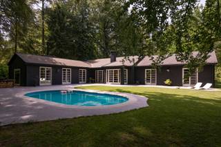 <p>Verscholen in het groen van Kapellenbos ligt deze karaktervolle gelijkvloerse villa op een bijzonder rustige en private locatie. De woning combineert een uitgesproken architectuur met warme materialen, veel licht en een sterke verbinding met de omliggende tuin en het bos.</p>
<p>Rond de woning werd een aangename buitenomgeving gecreëerd met een ruim terras, een ellipsvormig zwembad en mooie zichten op het groen. Dankzij de gunstige oriëntatie geniet de tuin vanaf de late voormiddag tot ’s avonds van de zon.</p>
<p>Ook binnen heeft de woning een heel eigen karakter. Donkere en warme houtsoorten, diepe kleuren en zorgvuldig gekozen materialen zorgen voor een gezellige en persoonlijke sfeer. De leefruimtes sluiten mooi op elkaar aan en bieden verschillende plekken om te ontspannen, te lezen of gasten te ontvangen.</p>
<p>De keuken werd uitgevoerd in inox en gecombineerd met sappeli-hout en een granito-betonvloer. Een doorkijkhaard zorgt voor extra verbinding en sfeer tussen de verschillende leefruimtes.</p>
<p>De master bedroom beschikt over een ruime maatwerkkastenwand en een eigen badkamer. De twee bijkomende slaapkamers bevinden zich in een aparte vleugel en delen een tweede badkamer, wat zorgt voor een aangename indeling en voldoende privacy.</p>
<p>Ook praktisch werd er goed nagedacht over de indeling. De ruime multifunctionele mudroom met onderhoudsvriendelijke vloer en maatkasten biedt plaats aan fietsen, sportmateriaal en alles wat je liever buiten de leefruimtes houdt.</p>
<p>Een bijzondere woning voor wie houdt van rust, privacy, groen en architectuur met karakter, op een van de aangenaamste locaties van Kapellenbos.</p>
<p>Grenzend aan het natuurdomein Klein Schietveld begint hier de stilte letterlijk aan de voordeur. Uren wandelen door uitgestrekte natuur zonder iemand tegen te komen wordt deel van het dagelijkse ritme. Dankzij de unieke hoekligging geniet het perceel van een uitzonderlijke privacy. De woning werd subtiel ingepland tussen indrukwekkende bomen en volwassen flora, waardoor ze haast verborgen opgaat in haar omgeving.</p>