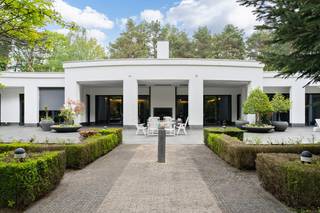 Rustig gelegen temidden van het groen,vindt u deze modernistische eenlaagse villa met een bewoonbare oppervlakte van maar liefst 900 m² op een mooi perceel van circa 10.000 m² voorzien van alle privacy. Via de imposante inkomhal met glazen dakconstructie betreden wij de villa met directe toegang tot de vestiaire, het gastentoilet en het kantoor. Centraal vindt u de woonkamer als één grote, open ruimte, badend in het licht met een prachtig uitzicht op de kleurrijke tuin. Links in de woonkamer bevindt zich de luxueus ingerichte bar, met daaraan grenzend het luxebinnenzwembad met wellnessgedeelte voor het ultieme vakantiegevoel. Door middel van grote raampartijen kan het zwembad indien gewenst afgesloten worden van de leefruimte. Naast het zwembad zijn er 2 ruime slaapkamers met ensuite dressing en badkamer. Rechts van de leefruimte treffen we de hedendaagse eilandkeuken voorzien van alle nodige comfort. Net als alle ruimtes heb je hier via grote schuiframen directe toegang tot het ruime verwarmde terras, dat zich uitstrekt over de volledige achtergevel. Door middel van screens kan het terras afgesloten worden waardoor je hier het hele jaar van kan genieten. De aanwezigheid van een houthaard en een buitendouche maakt het plaatje compleet. Net achter de keuken ligt de berging met bijkeuken. Hier bevinden zich zowel de lift als de trap naar de kelder, deze beslaat overigens dezelfde oppervlakte als de woning en bergt onder meer de technieken. De kelder is via een poort ook toegankelijk aan de zijkant van de woning door middel van een hellend gangpad, waardoor de immense oppervlakte zich perfect leent voorde opslag van goederen of desgewenst kan gebruikt worden als werkruimte. Verder leidt de berging naar de inpandige garage voor 2 wagens en de carport die nog eens ruimte biedt voor hetzelfde aantal wagens. Via een gang bereikt men de overige 2 slaapkamers, een extra badkamer en apart toilet. Deze gang beschikt over een aparte inkomdeur aan de zijkant van de woning. De tuin is volledig rondom het huis minutieus aangelegd en ZW gericht en vloeit naadloos over in het natuurpark “Mechelse Heide.” Verder is er ook een tuinhuis aanwezig. Het domein is volledig omheind en toegankelijk via een geautomatiseerde poort en beschikt over een beveiligingssysteem. Tot slot zijn er zonnepanelen aanwezig met een vermogen van 10200Wp. Deze uitgestrekte villa werd gebouwd met grote aandacht voor een perfecte integratie van de omliggende natuur. Kortom, wonen in een sterk staaltje architectuur, in een oase van groen. Luxe, comfort, rust en esthetiek gaan hand in hand in deze unieke eigendom die eveneens perfect geschikt is voor de uitoefening van een vrij beroep.<br /><br /><strong>Algemene Gegevens – Modernistische Villa</strong>
<ul>
	<li>Ligging: Rustig, midden in het groen</li>
	<li>Bewoonbare oppervlakte: ± 900 m²</li>
	<li>Perceeloppervlakte: ± 10.000 m²</li>
	<li>Oriëntatie tuin: Zuidwest (ZW)</li>
	<li>Volledige privacy</li>
</ul>
<br /><br /><strong>Indeling woning</strong>

<ul>
	<li>Inkomhal: Imposant, met glazen dakconstructie</li>
	<li>Vestiaire</li>
	<li>Gastentoilet</li>
	<li>Kantoor</li>
	<li>Woonkamer: Grote open leefruimte, veel licht, uitzicht op kleurrijke tuin</li>
	<li>Aansluitend: Luxueuze bar</li>
	<li>Binnenzwembad met wellnessgedeelte (afsluitbaar met glazen wanden)</li>
	<li>Keuken: Moderne eilandkeuken met alle comfort</li>
	<li>Directe toegang tot groot verwarmd terras over volledige achtergevel</li>
	<li>Terras afsluitbaar met screens, voorzien van houthaard en buitendouche</li>
	<li>Berging/bijkeuken:</li>
	<li>Lift en trap naar kelder</li>
	<li>Toegang tot inpandige garage (2 wagens) + carport (2 wagens)</li>
	<li>Slaapkamers:</li>
	<li>2 ruime slaapkamers met ensuite dressing en badkamer (naast zwembad)</li>
	<li>2 extra slaapkamers met extra badkamer en apart toilet</li>
	<li>Aparte zij-ingang via gang</li>
</ul>
<br /><br /><strong>Kelder</strong>

<ul>
	<li>Zelfde oppervlakte als woning (±900 m²)</li>
	<li>Toegankelijk via trap, lift en hellend gangpad</li>
	<li>Geschikt voor opslag of werkruimte</li>
	<li>Bevat technische installaties</li>
</ul>
<br /><br /><strong>Buitenruimte</strong>

<ul>
	<li>Professioneel aangelegde tuin rondom</li>
	<li>Naadloze overgang naar natuurpark “Mechelse Heide”</li>
	<li>Tuinhuis aanwezig</li>
	<li>Domein volledig omheind</li>
	<li>Automatische toegangspoort</li>
	<li>Beveiligingssysteem</li>
</ul>
<br /><br /><strong>Techniek &amp; Duurzaamheid</strong>

<ul>
	<li>Zonnepanelen: Vermogen 10.200 Wp</li>
	<li>Verwarming terras</li>
	<li>Lift aanwezig</li>
</ul>
<br /><br /><strong>Bijzonderheden</strong><br /><br />Deze modernistische villa combineert luxe, comfort en rust in een harmonisch geheel dat volledig opgaat in de omliggende natuur. De open architectuur, grote glaspartijen en hoogwaardige afwerking zorgen voor een licht en ruimtelijk gevoel.<br />De aanwezigheid van een binnenzwembad met wellness, een verwarmd terras, zonnepanelen en een lift bieden het hoogste wooncomfort. Dankzij de unieke ligging aan de rand van de Mechelse Heide geniet u van volledige privacy en een prachtig uitzicht.<br />Een uitzonderlijke eigendom waar architectuur en levenskwaliteit samenkomen – ideaal als exclusieve gezinswoning of voor de uitoefening van een vrij beroep.<br />- Maatregelenregister : Er zijn geen maatregelen op dit perceel<br /><br /><br /><strong>Locatie: Mechelen-aan-de-Maas, Maasmechelen</strong><br /><br />Rustig gelegen doch centraal. Faciliteiten zoals scholen, winkels en diensten vindt u in de nabijheid. Als kers op de taart geniet u een vlotte verbinding met invalswegen naar Brussel Antwerpen en Nederland.