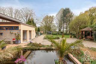Zeer goed onderhouden architectenwoning op een toplocatie met uitvalbasis naar alle voorzieningen, winkels en aansluiting met hoofdwegen. Daarentegen geniet u achteraan van een rustig, open en groen uitzicht wat zorgt voor een oase van rust en privacy. De woning beschikt over een gunstig EPC-label C, dubbele beglazing, alarm, een nieuwe condenserende ketel op stookolie van 2022, lucht-lucht warmtepompen in verschillende ruimtes en een bruikbare vloeroppervlakte van maar liefst 315m2.<br /><br />Indeling:<br />Via de voorzijde van de woning betreedt u de inkomhal, die toegang geeft tot het architectenatelier, het bureel, een opslagruimte en een gastentoilet. Via de rechterzijde komt u in de privé-inkomhal, vanwaar u de lichtrijke leefruimte bereikt met zicht op de tuin, evenals de volledig geïnstalleerde keuken. Heel de woning is afgewerkt met hoogwaardige materialen. Zowel het werk- als het privégedeelte zijn intern met elkaar verbonden. Op de eerste verdieping geeft de nachthal toegang tot 4 volwaardige slaapkamers. Verder vindt u hier een badkamer en een apart toilet. Daarnaast beschikt de woning over een kelder. Het pand is uitermate geschikt voor meerdere commerciële doeleinden, kangoeroe woning...<br /><br />Buiten beschikt de woning over een overdekt terras met vijver, evenals een ruime oprit die doorloopt tot achteraan, met toegang tot de praktische garages en opslagruimtes. De tuin is zuidgericht en achteraan bevindt zich een wandelpad, vanwaar ook toegang wordt geboden tot de tuin. <br /><br />G-score: A – P-score: A. <br />Voor info of bezichtiging: 054 23 53 83 of info@localsvastgoed.be