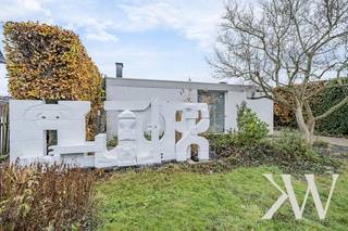 <em>Wie Katrien Wouters zegt, zegt vastgoed met een tikkeltje meer. En dat is niet zomaar.<br /><br />Deze brutalistisch-modernistische woning belichaamt het samenspel van architectonische finesse en eigentijdse leefkwaliteit. Het ontwerp is afkomstig van de hand van architect Adolf Nivelle, in co-creatie met de bouwheer, kunstenaar Toon Vanmechelen.</em><br /><br />Hier geen overdaad: zichtbaar beton, baksteen en hout tonen eerlijk hun structuur. Deze architectuur sluit aan bij een internationale beweging die eenvoud en functionaliteit benadrukt. Elk detail is doordacht, elke overgang zorgvuldig georkestreerd, met als resultaat een leefomgeving die niet alleen esthetisch intrigeert, maar ook functioneel tot in de puntjes klopt.<br /><br /><strong>Architecturaal geheel</strong><br />De woning presenteert zich als een monument van ingetogen kracht. Aan de straatzijde is het huis omhuld in privacy zonder het licht volledig buiten te sluiten. De witgeschilderde bakstenen gevel is strak, maar niet steriel. De geometrie is helder, de compositie evenwichtig, en het geheel wordt verankerd in een groene context die niet louter decoratief is, maar deel uitmaakt van de beleving.<br />Naar achteren toe opent de woning zich volledig. Grote raampartijen creëren een genereuze relatie met de tuin. De architectuur zoekt hier geen spektakel, maar rust en structuur – een helder kader voor het dagelijkse leven.<br /><br /><strong>Ruimtelijke helderheid en materiële rust</strong><br />De woning is opgebouwd volgens een doordachte structuur van beton, baksteen, glas en staal. Grote raampartijen verbinden binnen en buiten, terwijl horizontale lijnen ritme en stabiliteit brengen. Het resultaat is een leefomgeving die tegelijk open en geborgen aanvoelt. <br />Nivelle's architectuur staat in het teken van helderheid. Het gebruik van ruwe materialen, geometrische vormen en strakke lijnen zorgt voor een ingetogen maar krachtige uitstraling en geeft de woning een tijdloos karakter. <br /><br /><strong>Leefruimte in overvloed</strong><br />Binnenin ontvouwt de woning zich in een sequentie van ruimten die logisch op elkaar aansluiten, maar toch hun eigen sfeer behouden. De leefruimte is breed en licht, met warme houten vloeren, grote raampartijen en een ingebouwde kastenwand die zorgt voor rust en overzicht. Kunst krijgt een prominente plaats in het interieur en ook de koperen houthaard is een echte eyecatcher. De keuken is tegelijk functioneel en esthetisch: een strakke, heldere ruimte met een praktische opstelling. Op het gelijkvloers vind je daarnaast 4 slaapkamers, een badkamer, kunstatelier en een inpandige garage met geautomatiseerde sectionaalpoort. <br />                                                                                                                       <br /><strong>Integratie van kunstwerken</strong><br />De beeldhouwwerken in de voortuin en patio fungeren als symbool en verlengstuk van de architectuur. Kunst en gebouw zijn onlosmakelijk met elkaar verbonden. Meer dan een huis is dit een creatieve biotoop. Door zijn integrale ontwerpvisie creëerde de architect een omgeving die inspireert. <br /><br /><strong>Architectuur met toekomstpotentieel</strong><br />Deze woning is niet louter een object uit het verleden, maar biedt ook perspectief. Hoewel er energetisch werk aan de winkel is — het EPC-label F vraagt om renovatie — blijft de structuur bijzonder robuust. Dit is een pand dat vraagt om een eigentijdse interpretatie, zonder het originele karakter uit te wissen. Voor wie houdt van architectuur met ziel en geschiedenis, biedt deze woning een zeldzame kans.<br /><br /><strong>Meer ontdekken?</strong><br />Wil je graag thuiskomen in deze uitzonderlijke woning?<br />Contacteer mij dan snel voor meer info of een persoonlijk bezoek ter plaatse!<br /><br /><em>De vermelde oppervlaktes zijn slechts indicatief. Katrien Wouters - creating IMMOtions kan niet verantwoordelijk gesteld worden voor de juistheid van de aan haar verstrekte gegevens.  </em>