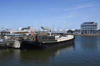 Bekijk deze boot uitgebreid op www.koosi.beDeze prachtige woonboot ‘Ons Genoegen’ is een blikken klipper van 1893 die ooit gebouwd is om steenkool te vervoeren over het water. De boot is gelegen op de mooiste plaats aan het Houtdok-Zuidkaai, de instapprijs van de concessie is inbegrepen in de koopsom. De romp is gemaakt uit staal van 6 mm dik en is de laatste keer officieel gecontroleerd in oktober 2024. Via de aanlegsteiger betreden we de boot en komen we op het achterdek waar de stuurinrichting van de boot is geplaatst. Als we dan binnenkomen in de leefruimte voelen we direct de gezelligheid die deze woonboot kan bieden, zeker door de aanwezigheid van de speksteenkachel. Deze ruimte is zeer groot en door de leuke lichtinval wordt dit nog eens extra benadrukt. Op het hogere deel is momenteel het salon geplaatst vanwaar je door de vele ramen direct zicht hebt op het water. Als we dan een paar trappen naar beneden lopen komen we in het gedeelte waarin de zeer ruime en volledig ingerichte keuken is gebouwd. Aan de andere kant vinden we de grote eettafel en toegang tot de ‘kelderruimte’. In voorste deel van boot zijn de slaapvertrekken voorzien. Vanuit de leefruimte komen we in een gang met rechts een berging waar wasmachine en droogkast kunnen geplaatst worden. Aan de overzijde van deze berging vinden we de technische ruimte met verwarmingsketel en pompen. Naast deze technische ruimte is het toilet gelegen. Iets verder in de gang vinden we rechts (stuurboordzijde) een slaapkamer met dubbel bed. Over deze slaapkamer (bakboordzijde) is de eerste badkamer met douche voorzien. Nog iets verder rechts is dan de 2de slaapkamer voorzien, deze kamer is momenteel ingericht als dressing en heeft een enkel bed. Over deze slaapkamer ligt de 2de badkamer, ook voorzien met lavabo en douche. Helemaal vooraan de boot komen we in de grote slaapkamer met ingebouwde kasten.