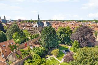 Binnen het Karmelproject bevindt zich een van de meest bijzondere woonentiteiten van het domein: de Kapelloft. Gevestigd in de voormalige neogotische kloosterkapel uit 1877, biedt deze eigendom een woonervaring die zelden voorkomt op de vastgoedmarkt.<br /><br />Bij de herbestemming van de kapel werd gekozen voor een doordacht box-in-the-box-concept, waarbij de oorspronkelijke kapelstructuur maximaal behouden blijft. Hierdoor blijven de indrukwekkende volumes, de hoge gewelven, de spitsboogvensters en het historische karakter van de ruimte volledig beleefbaar.<br /><br />De leefruimte bevindt zich centraal in de kapel en wordt gekenmerkt door een uitzonderlijk ruimtegevoel. Vanuit de keuken biedt een groot raam zicht op de uitgestrekte parktuin van het domein. Een draaitrap leidt naar een hoger gelegen bureau- of polyvalente ruimte die uitkijkt over het geheel.<br /><br />Aan de andere zijde van de loft bevindt zich een afzonderlijke woonmodule. Op het gelijkvloers zijn de master bedroom en de ensuite badkamer ondergebracht. Op de verdieping bevinden zich nog twee bijkomende slaapkamers en een tweede badkamer.<br /><br />Naast de privatieve woonruimtes genieten bewoners van toegang tot de gemeenschappelijke kloostertuin van circa 5.000 m². Met haar historische elementen, wandelpaden en volwassen groen vormt deze tuin een uitzonderlijke plek van rust midden in het historische centrum van Brugge.<br /><br />De Kapelloft toont hoe erfgoed een nieuwe bestemming kan krijgen zonder zijn identiteit te verliezen. Een eigendom waar geschiedenis, architectuur en hedendaags wooncomfort samenkomen in een setting die nauwelijks een vergelijking kent.<br /><br />Een unieke woonkans voor wie op zoek is naar ruimte, karakter en rust op een van de meest bijzondere locaties van Brugge.