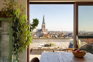 <strong>Algemeen</strong><br />Op de zesde en zevende verdieping, helemaal bovenaan het gebouw, ligt dit duplex-penthouse van ca. 211 m². Het voelt eerder als een huis in de lucht dan als een appartement. Beneden is het rustig, met drie slaapkamers, een aparte dressing, een badkamer en veel ingemaakte kasten. Boven ligt één grote, lichte leefruimte met witgeschilderde balken, een industriële keuken, een vierde slaapkamer, een tweede badkamer en twee terrassen van samen zo’n 70 m². Een smeedijzeren wenteltrap verbindt de twee verdiepingen. Ook technisch zit het goed: het dak van het gebouw werd gerenoveerd, de verwarmingsketel is pas vernieuwd en het appartement heeft een EPC-label C. Er is een lift, en bij het appartement horen twee kelders.<br /><strong>Ligging</strong><br />Het penthouse ligt aan het begin van de Turnhoutsebaan, op het kruispunt met de Carnotstraat en de Kerkstraat. Het Centraal Station, het Astridplein en de Zoo liggen op een paar minuten wandelen. Winkels, bakkers, koffiebars en restaurants vind je in alle richtingen, en tram en bus stoppen vlakbij. Je woont hier in het hart van de stad, maar vanaf de bovenste verdieping hoor je de drukte nauwelijks en zie je vooral daken, kerktorens en veel lucht.<br /><strong>Indeling</strong><br />Zesde verdieping: de nachtverdieping<br />Je komt binnen in een ruime inkomhal met een marmeren vloer en een hele wand ingemaakte kasten, zodat jassen, schoenen en tassen meteen uit het zicht zijn. Vanuit de hal bereik je alle ruimtes op deze verdieping.<br />Op deze verdieping liggen drie slaapkamers. De grootste is een echt ruime kamer met een groot raam en een deur naar het balkon, vanwaar je over de daken en het groen van de buurt kijkt. De tweede slaapkamer is nu ingericht als bureau. Een groot raam zorgt voor veel daglicht en de witgeschilderde bakstenen muur geeft de kamer karakter. De derde slaapkamer is even rustig en ruim, en elke kamer heeft eigen kastruimte. Daarnaast is er een aparte dressing met een eigen raam, dat uitkijkt op de kerktoren. Zo heb je kleding en schoenen netjes uit de slaapkamers. Op de hele verdieping, behalve in de inkomhal, ligt eiken mozaïekparket. De korte stroken liggen per vier in vierkantjes, telkens een kwartslag gedraaid zoals een dambord. Het is een klassieker die mooi verweerd is en de kamers meteen warmte geeft.<br />De badkamer op deze verdieping is sober en strak. De douche heeft grote antracietkleurige tegels, een regendouche met handdouche en een glazen schuifdeur. Het aparte toilet ligt ernaast.<br />De wenteltrap<br />Vanuit de inkomhal loop je via een smeedijzeren wenteltrap naar boven. Wie omhoog kijkt, ziet een ronde trapopening met een sierlijke balustrade vol krullen en daarboven de witte balken van de leefruimte. Het is een van die details die je nergens anders tegenkomt.<br />Zevende verdieping: de leefverdieping<br />Boven kom je uit in het hart van de woning: één grote leefruimte van meer dan twaalf meter lang. Het plafond heeft witgeschilderde houten balken en aan meerdere kanten zijn er ramen, waardoor er van ‘s ochtends tot ‘s avonds licht binnenvalt. De ronde trapopening staat midden in de ruimte en verdeelt ze vanzelf in zones zonder dat er muren nodig zijn. Aan de ene kant is er plaats voor een grote zithoek, met een witte nis in de muur en een stuk witgeschilderde baksteen die er een gezellige hoek van maken. Aan de andere kant staat de eettafel voor een groot raam met houten schuiframen, die rechtstreeks uitkomen op het terras.<br />De keuken is industrieel ingericht: witte metrotegels tot aan het plafond, een stoer professioneel fornuis met grote oven, een roestvrijstalen dampkap met zichtbare afvoerbuis, open rekken in staal en hout en een magneetstrip voor de messen. De dubbele spoelbak staat onder een breed raam, zodat je bij de afwas over de stad kijkt. Het is een keuken voor mensen die graag en veel koken.<br />Dan de terrassen. Het zuidwestterras van ca. 40 m² krijgt de zon in de namiddag en ‘s avonds. Het noordoostterras van ca. 30 m² is ideaal voor een ontbijt in de ochtendzon of als koele plek op warme zomerdagen. Op het terras liggen terrazzotegels en rondom staat een witgeschilderde bakstenen borstwering. Het uitzicht reikt over de daken tot aan de skyline van Antwerpen.<br />Op deze verdieping ligt ook de vierde slaapkamer, een rustige kamer met sierlijsten aan het plafond. De tweede badkamer is een echte eyecatcher. De wanden zijn bekleed met handgemaakte, glanzende tegeltjes in zachte grijsblauwe tinten, en er is een ligbad, een inloopdouche met regendouche, een lange brede wastafel met een grote spiegelkast en een handdoekradiator. Door het raam valt daglicht binnen. Verder is er een apart toilet en een berging die als wasplaats is ingericht.<br />De hele bovenverdieping heeft een bamboevloer met de typische knopen in het hout, die een ander karakter heeft dan het parket beneden. In de leefruimte zijn de vloerconvectoren weggewerkt onder houten lattenroosters, zodat er geen radiatoren tegen de muren staan.<br /><strong>Troeven</strong>
<ul>
<li>Duplex-penthouse van ca. 211 m² op de 6e en 7e verdieping</li>
<li>Twee terrassen: zuidwest (ca. 40 m²) en noordoost (ca. 30 m²), met weids zicht over Antwerpen</li>
<li>Leefruimte van ruim 2 m1 met witgeschilderde balken en veel lichtinval</li>
<li>Industriële keuken met professioneel fornuis</li>
<li>4 ruime slaapkamers (3 op de 6e, 1 op de 7e), een dressing en veel ingemaakte kasten</li>
<li>2 volwaardige badkamers (één per verdieping) en 2 aparte toiletten</li>
<li>Smeedijzeren wenteltrap als blikvanger</li>
<li>Eiken mozaïekparket op de 6e verdieping, bamboe op de 7e, marmer in de inkomhal</li>
<li>Vloerconvectoren in de leefruimte</li>
<li>Gerenoveerd dak, nieuwe verwarmingsketel, EPC C</li>
<li>Lift en 2 kelders</li>
<li>Op wandelafstand van het Centraal Station</li>
</ul>
De grondplannen, het EPC en de asbestinventaris vind je onder “Documenten” op onze website. Kom het uitzicht zelf bekijken en maak een afspraak via info@wellimmo.net of op 03 225 00 68.