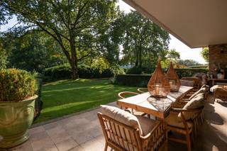 <p>Dit uitzonderlijk ruime en instapklare gelijkvloers appartement combineert een royale woonoppervlakte met een hoogwaardige afwerking, een aangename lichtinval en een doordachte indeling. Met ca. 200 m² bewoonbare oppervlakte, een ruim en zonnig terras en rechtstreekse toegang tot de prachtig aangelegde parktuin met vijver, biedt deze unieke eigendom een bijzonder aangename woonbeleving.</p>
<p>Het appartement beschikt over drie volwaardige slaapkamers, twee badkamers, een aparte wasplaats en bijkomende bergruimte in de kelder. Daarnaast zijn er twee ruime parkeerplaatsen in de ondergrondse garage.</p>
<p><strong>Indeling</strong></p>
<p>Via de stijlvolle en lichtrijke gemeenschappelijke inkomhal bereikt u het appartement. De centrale inkomhal beschikt over een vestiaire en gastentoilet en biedt toegang tot zowel de leefruimtes als de keuken.</p>
<p>De luxueus afgewerkte keuken is uitgerust met kwalitatieve inbouwtoestellen, waaronder een vaatwasser, koelkast, diepvriezer, combi-oven en stoomoven.</p>
<p>De bijzonder ruime leefruimte is afgewerkt met tijdloze eiken parket en omvat een sfeervolle eethoek, een gezellige tv-hoek en een aparte leesruimte. Dankzij de grote raampartijen geniet de ruimte van een overvloed aan natuurlijk licht en een prachtig zicht op de aangelegde tuin en vijver. Vanuit de leefruimte is er rechtstreeks toegang tot het ruime, zonnige terras.</p>
<p>Via de nachthal bereikt u de drie volwaardige slaapkamers. Eén van de kamers is momenteel ingericht als bureau en beschikt over een aansluitende wasplaats. De twee overige slaapkamers beschikken elk over een privatieve buitenruimte en sluiten aan op respectievelijk een douchekamer en een badkamer.</p>
<p><strong>Bijzonderheden</strong></p>
<ul>
<li>
<p>Instapklaar en luxueus afgewerkt gelijkvloers appartement van ca. 200 m²</p>
</li>
<li>
<p>Ruim en zonnig terras met zicht op de prachtig aangelegde parktuin en vijver</p>
</li>
<li>
<p>Drie volwaardige slaapkamers</p>
</li>
<li>
<p>Twee badkamers</p>
</li>
<li>
<p>Volledig voorzien van kwalitatief maatwerk en ingemaakte kasten</p>
</li>
<li>
<p>Hoogwaardige afwerking met onder meer eiken parket en natuursteen</p>
</li>
<li>
<p>Uitstekende beveiliging met alarmsysteem</p>
</li>
<li>
<p>Twee ruime parkeerplaatsen in de ondergrondse garage</p>
</li>
<li>
<p>Extra bergruimte in de kelder</p>
</li>
</ul>