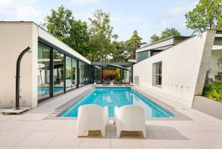 Op een absolute toplocatie in het groene Poppel, verscholen in een rustige residentiële omgeving en grenzend aan de natuur, bevindt zich deze uitzonderlijke architecturale villa. Een eigendom waar luxe, privacy, ruimte en verfijning op harmonieuze wijze samenkomen. Dankzij de doordachte architectuur, hoogwaardige materialen en de naadloze verbinding tussen binnen- en buitenruimtes geniet u hier elke dag van een exclusieve woonbeleving.<br /><br />Bij het betreden van de woning wordt u verwelkomd in een inkomhal met vestiaire en een apart gastentoilet. Vanuit deze royale entree bereikt u zowel de leefruimtes als, via een stijlvolle glazen doorgang, de afzonderlijke slaapvleugel.<br /><br />De bijzonder lichtrijke leefruimte vormt het hart van de woning. Grote glaspartijen brengen het omliggende groen naar binnen en creëren een unieke interactie tussen de woning, de Japanse binnentuin en het zwembad. De sfeervolle gashaard zorgt voor warmte en gezelligheid, terwijl één van de raampartijen is uitgevoerd in innovatief schakelbaar glas voor extra comfort op zonnige dagen.<br /><br />De hypermoderne leefkeuken werd in 2023 verder geoptimaliseerd met een Liebherr Fresh-koelkast, Siemens inductiekookplaat en combi-oven, een Miele vaatwasser en een Quooker. De maatwerkkasten en de praktische bijkeuken maken het geheel compleet.<br /><br />Ook het souterrain werd ingericht met oog voor ontspanning en functionaliteit. Hier bevinden zich een ruime homecinema, een volledig ingerichte fitnessruimte, een technische ruimte en een royale inpandige garage voor twee wagens. Indien gewenst kan de fitnessruimte opnieuw bij de garage worden betrokken, waardoor plaats ontstaat voor een derde wagen. Daarnaast beschikt de woning over parkeergelegenheid voor maar liefst negen bezoekerswagens.<br /><br />Via de volledig beglaasde doorgang, met zicht op de sfeervol verlichte Japanse binnentuin, bereikt u de slaapvleugel. De master suite beschikt over een royale slaapkamer en een smaakvol vernieuwde badkamer (2019), uitgerust met een ligbad, dubbele wastafel en een exclusieve Sunshower met keuze tussen infraroodwarmte of UV-licht.<br /><br />Daarnaast zijn er nog twee ruime slaapkamers en een tweede badkamer, die in 2025 volledig werd vernieuwd. De aansluitende wellnessruimte beschikt over een sauna voor zes personen met zowel een traditionele saunakachel als een infraroodfunctie. Een praktische wasplaats vervolledigt deze vleugel.<br /><br />Ook buiten werd werkelijk aan alles gedacht. Het ruime overdekte terras vormt een natuurlijk verlengstuk van de leefruimte en kijkt uit over de prachtig aangelegde tuin en het verwarmde zwembad. Dankzij het vernieuwde solardek warmt het zwembadwater tijdens zonnige dagen op tot ongeveer 29,5°C, terwijl een automatische zwembadrobot instaat voor het onderhoud.<br /><br />De Japanse binnentuin vormt het architecturale middelpunt van de woning en wordt 's avonds sfeervol verlicht. Dankzij een geïntegreerd drainagesysteem en een automatische druppelirrigatie blijft deze tuin het hele jaar door perfect onderhouden. Ook de voor- en achtertuin beschikken over een geautomatiseerd RainBird-beregeningssysteem met WiFi-bediening, gevoed via een geslagen waterput.<br /><br />Achteraan op het perceel, discreet verscholen tussen het groen maar volledig op eigen terrein, bevindt zich een tuinhuis van ca. 18 m², voorzien van elektriciteit en verlichting.<br /><br />Ook op technisch vlak voldoet deze villa aan de hoogste verwachtingen. Zo beschikt de woning over 28 zonnepanelen (11 kW), een Alfen Pro-laadpaal, automatische screens aan de glazen gang, master bedroom en het overdekte terras, een professioneel alarm- en camerabewakingssysteem en een gunstig EPC-label B.<br /><br />Deze uitzonderlijke villa biedt een zeldzame combinatie van architectuur, luxe, comfort, technologie en privacy op een unieke locatie midden in het groen. Een exclusieve woonbeleving voor wie enkel genoegen neemt met het allerbeste.<br /><br />Bijzonderheden<br />*Absolute topligging in een rustige, residentiële omgeving grenzend aan de natuur<br />*Vernieuwde leefkeuken (2023) met hoogwaardige Siemens-, Miele-, Liebherr- en Quooker-uitrusting<br />*Homecinema, fitnessruimte en inpandige garage voor 2 à 3 wagens<br />*Luxueuze masterbedroom met een ensuite badkamer, uitgerust met een Sunshower.<br />*Tweede badkamer volledig vernieuwd in 2025<br />*Wellnessruimte met sauna voor 6 personen (traditioneel & infrarood)<br />*Verwarmd zwembad met vernieuwd solardek en automatische zwembadrobot<br />*Geautomatiseerde screens en RainBird-beregeningssysteem<br />*Professioneel alarm- en camerabewakingssysteem<br />*28 zonnepanelen (11 kW) en Alfen Pro-laadpaal<br /><br />Uitstekende verbinding richting Nederland en omliggende gemeenten.