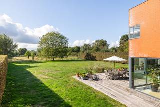 Welkom in de Lovelddreef, een unieke kans om riant en instapklaar te wonen in het felbegeerde, residentiële Heusden. Deze vrijstaande villa (bouwjaar 1993) onderscheidt zich door haar opvallende architectuur naar de hand van arch. Fritz Schaffrath in warme rode baksteen en bevindt zich op een indrukwekkend perceel van 1.788 m². Hier vloeien doordacht design, leisteen accenten, overvloedige lichtinval en weidse natuur naadloos in elkaar over.<br /><br />Bij het betreden van de woning via de stijlvolle inkomhal valt meteen de royale bruikbare oppervlakte van 263 m² op. De leefruimtes worden getypeerd door een speelse split-level indeling, plafondafwerking in karaktervol zichtbeton en indrukwekkende glazen raampartijen van vloer tot plafond. De sfeervolle zithoek is voorzien van een strakke open haard en parketvloer, terwijl de verhoogde eetruimte is ondergebracht in een spectaculaire glazen uitbouw met panoramisch zicht op de tuin en achterliggende velden. Om het hele jaar door van een optimaal binnenklimaat te genieten, is de achtergevel voorzien van hoogwaardig zonnewerend glas.<br /><br />Via de glazen schuifdeuren stapt u zo het riante houten terras op, de perfecte plek voor uitgebreid tafelen en ontspannen in alle rust. De omheinde, groene tuin met karaktervolle volwassen bomen garandeert 100% privacy en biedt een veilige speelomgeving voor kinderen.<br /><br />De strakke, moderne keuken staat in directe verbinding met de leefruimte en is voorzien van speelse doorkijkjes naar het groen. Via de stijlvolle traphal bereikt u de lichtrijke overloop op de eerste verdieping, die prachtig wordt verlicht door een natuurlijke lichttunnel.<br /><br />Op de bovenverdieping bevinden zich vier volwaardige slaapkamers en twee badkamers. Bijzonder troefrijk voor gezinnen zijn de twee aaneengeschakelde kinderkamers die rechtstreeks met elkaar verbonden zijn via een schuifdeur. Dit biedt kinderen of tieners de unieke mogelijkheid om hun slaapkamer direct te combineren met een eigen speel-, studeer- of ontspanningsruimte. Een derde, afzonderlijke (kinder)kamer vervolledigt het plaatje en biedt heel wat mogelijkheden. De master suite is eveneens een blikvanger: met zijn royale oppervlakte, zicht op het groen en met een adembenemende en-suite badkamer waar het ligbad pal voor een groot raam is geplaatst, met direct zicht op de tuin en de boomtoppen. Elke slaapkamer is voldoende ruim om dubbele bedden te plaatsen.<br />Aan comfort en functionaliteit is tot in de puntjes gedacht: een ruime oprit met een brede dubbele carport, een handige buitenberging, een droge voorraadkelder, een waterontharder en een geïntegreerde surround installatie.<br /><br />Kortom, hier combineert men architecturale klasse en instapklaar gezinscomfort met een droomperceel in het groen. We nodigen u van harte uit voor een individueel en persoonlijk begeleid bezoek.<br /><br />Belangrijk: de vermelde prijs is een richtprijs!<br /> 