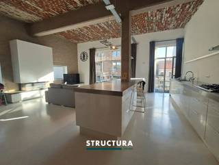 In het centrum van Merchtem bevindt zich deze ruime loft met een bewoonbare oppervlakte van ca. 133 m², ondergebracht in de voormalige Azijnbrouwerij. Het industriële karakter van het gebouw is mooi bewaard gebleven en komt onder meer tot uiting in de hoge plafonds, de authentieke plafondstructuur en de grote raampartijen.De leefruimte vormt het hart van de loft en combineert een open karakter met veel natuurlijke lichtinval. De gashaard zorgt voor extra sfeer en sluit mooi aan bij de industriële uitstraling van het geheel. Met drie slaapkamers, een EPC-label B en een centrale ligging biedt deze loft bovendien een interessante combinatie van ruimte, karakter en energiezuinigheid.IndelingEerste verdieping: Inkomhal met apart toilet, ruime en lichtrijke leefruimte, open keuken, terras, drie slaapkamers, douchekamer en badkamer met zowel bad als douche. Gelijkvloers: garage, bij aan te kopen aan €25.000 Karakteristieken- EPC-label B- Momenteel verhuurd aan € 1.175/maand- Centrale ligging in Merchtem- Zuid georiënteerd terrasEen karaktervolle loft waar de geschiedenis van het gebouw nog duidelijk voelbaar is, gecombineerd met een ruime indeling en hedendaags wooncomfort. Dankzij de drie slaapkamers, de oppervlakte van ca. 133 m² en het gunstige EPC-label B is dit een interessante eigendom voor wie op zoek is naar een ruime loft in het centrum van Merchtem. Contacteer ons via 02 460 80 46 of info@structura.be