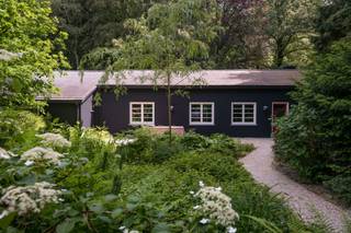 <p>Verborgen in het groen van Kapellenbos, midden in één van de meest bosrijke en rustige stukken van Antwerpen, ligt een plek die zelden op de markt verschijnt. Geen klassieke woning, maar een huis met een ziel, ontworpen vanuit gevoel, rust en verbondenheid met de natuur.</p>
<p>Wat deze plek extra bijzonder maakt, is het licht. In een omgeving waar hoge bomen vaak het zonlicht filteren, werd hier een zeldzame balans gevonden: vanaf de late voormiddag glijdt de zon doorheen de tuin en blijft ze aanwezig tot het laatste avondlicht. Elk moment van de dag brengt een andere sfeer.</p>
<p>Aan het leefterras werd een granito-betonterras gegoten dat op een natuurlijke manier contrasteert met het iconische mid-century ellipsvormige zwembad. Daarachter ontvouwt zich een adembenemend zicht op het omliggende beukenbos, een decor dat elk seizoen een ander karakter krijgt.</p>
<p>De woning werd ontworpen door twee creatieve persoonlijkheden met één duidelijke visie: een huis creëren dat zowel als zomer- én winterhuis perfect aanvoelt. Een plek waar gezelligheid, warmte en leefbaarheid altijd primeren. De zwarte laagbouwvilla, met haar uitgesproken Hamptons-geïnspireerde architectuur en verfijnde witte art-deco raamverdelingen, fonkelt subtiel tussen het groen.</p>
<p>Binnen werd gekozen voor rijke, warme materialen en zorgvuldig opgebouwde contrasten. Japanse roesttinten zorgen onmiddellijk voor een omarmend gevoel, een interieur waarin je vanzelf vertraagt en wil blijven hangen.</p>
<p>De leefruimte werd ontworpen als een plek van rust, muziek, gesprekken en creativiteit. De volledig geïntegreerde bibliotheek in sappeli-hout, gecombineerd met een energiezuinige open haardcassette in geborstelde inox, werd op maat ontworpen door designstudio Altu Space. Samen met het diepe groene tapijt ontstaat hier een sfeer die tegelijk luxueus en ingetogen aanvoelt.</p>
<p>De eigenaars, liefhebbers van het bredere modernisme, trokken die visie consequent door in de volledige woning. Typische Berker-schakelaars, warme houtaccenten en verfijnde materialiteit versterken de subtiele “cabin in the woods”-sfeer. Ook de keuken ademt karakter: een krachtige combinatie van een granito-betonvloer, een volledig inox Bossuyt-keuken en sappeli-houten wand- en plafondafwerking. De doorkijkhaard verbindt de ruimtes op sfeervolle wijze en maakt van winterdagen iets uitzonderlijk gezelligs.</p>
<p>De badkamer werd tailor made gerealiseerd in epoxy. De eigenaars maakten het nog aparter. Door de spiegelwanden wordt er een cinematografische ruimte gecreëerd die tegelijk minimalistisch en toch bijzonder warm aanvoelt. In de master bedroom keert het rijke houtgebruik terug in de volledige maatwerkkastenwand, wat de ruimte een serene hotelachtige rust geeft.</p>
<p>De woning werd bovendien intelligent ingedeeld: de twee extra slaapkamers bevinden zich in een aparte vleugel met eigen badkamer, ideaal voor kinderen of gasten. Ook hier keren de warme houtaccenten en zachte, gebogen lijnen terug.</p>
<p>Tot slot beschikt de woning over een bijzonder praktische en tegelijk zeldzame “mudroom”: een ruimte ontworpen voor wie houdt van wandelen, sporten en buitenleven. Met PU-vloeren en inox maatkasten werd deze zone bewust ontworpen om zonder zorgen modderige schoenen, fietsen of outdooractiviteiten op te vangen, zonder in te boeten aan esthetiek.</p>
<p>Dit is geen huis dat zich laat vergelijken. Het is een plek die je voelt. Een woning waar architectuur, natuur en rust op een uitzonderlijke manier samenvallen.</p>
<p>Zulke plekken komen zelden te koop. Misschien maar één keer.</p>